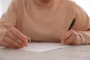 Picture of a woman signing a paper on the table in front of her while holding her wedding ring in her other hand.