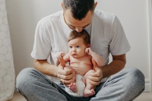 Picture of a father sitting cross-legged on the ground, holding a baby in his lap and kissing the baby's head.