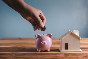Picture of a person placing a coin into a piggy bank next to a miniature wooden house figurine.