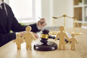Picture of wooden figurines of a mother, father, and two children surrounding a gavel, with an attorney and the scales of justice in the background.