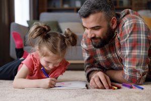 Picture of a father and his young daughter lying on the floor and coloring with crayons.
